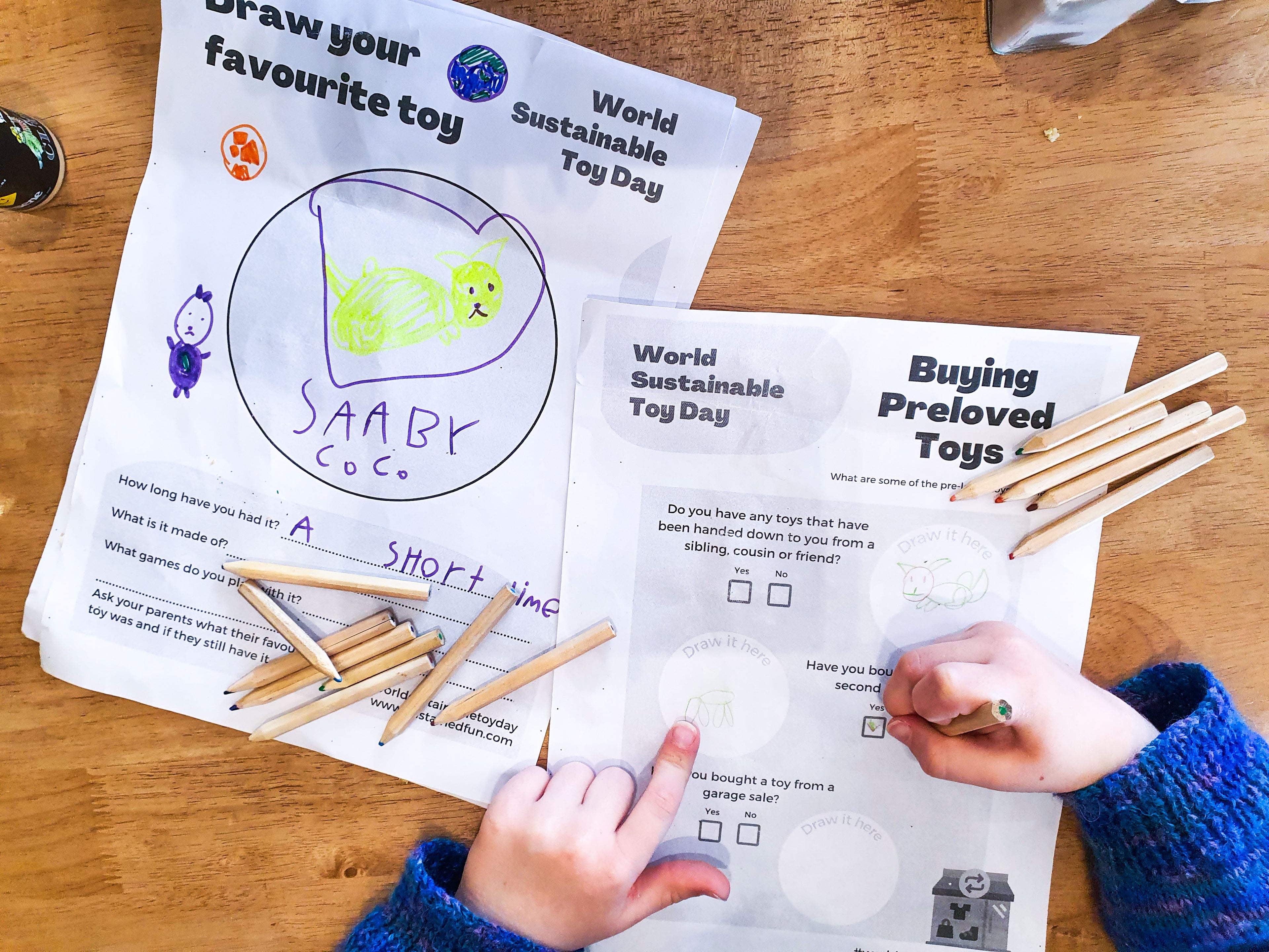 A bird's-eye view of activity sheets with coloured pencils and a young girl's hands. The young girl is completed one of the activity sheets. The sheets say: 'Draw your favourite toy, World Sustainable Toy Day' and 'Buying pre-loved toys. World Sustainable Toy Day'.