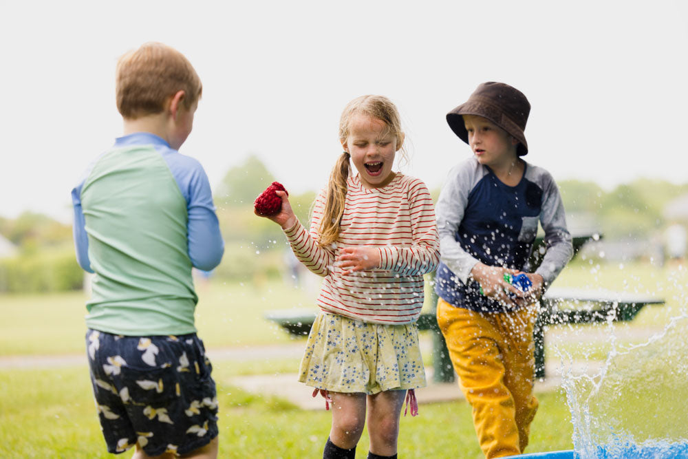 Three 10 year old children playing with EcoSplat reusable water balloons in the park. There are two boys and one girl. Water is dripping off the water balloons, they are laughing.