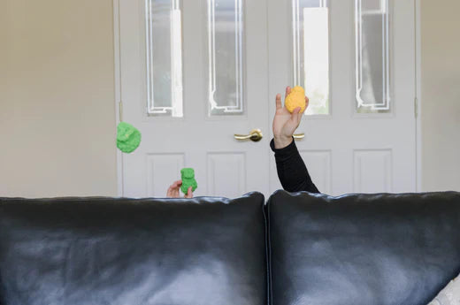 A black sofa in a living room, there are two people hiding behind the sofa throwing EcoSplat reusable water balloons over the back of the sofa. All you can see are their arms above the back of the sofa