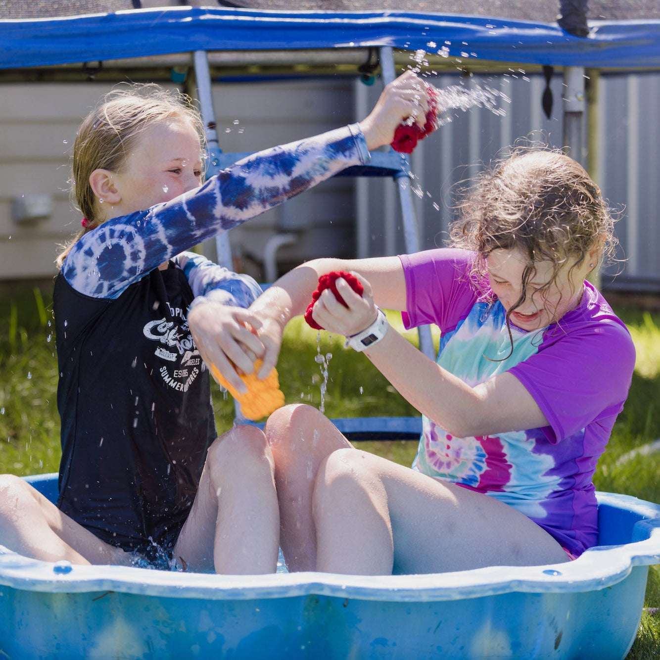 Two girls laughing and sitting in a paddling pool and playing with EcoSplat Reusable Water Balloons