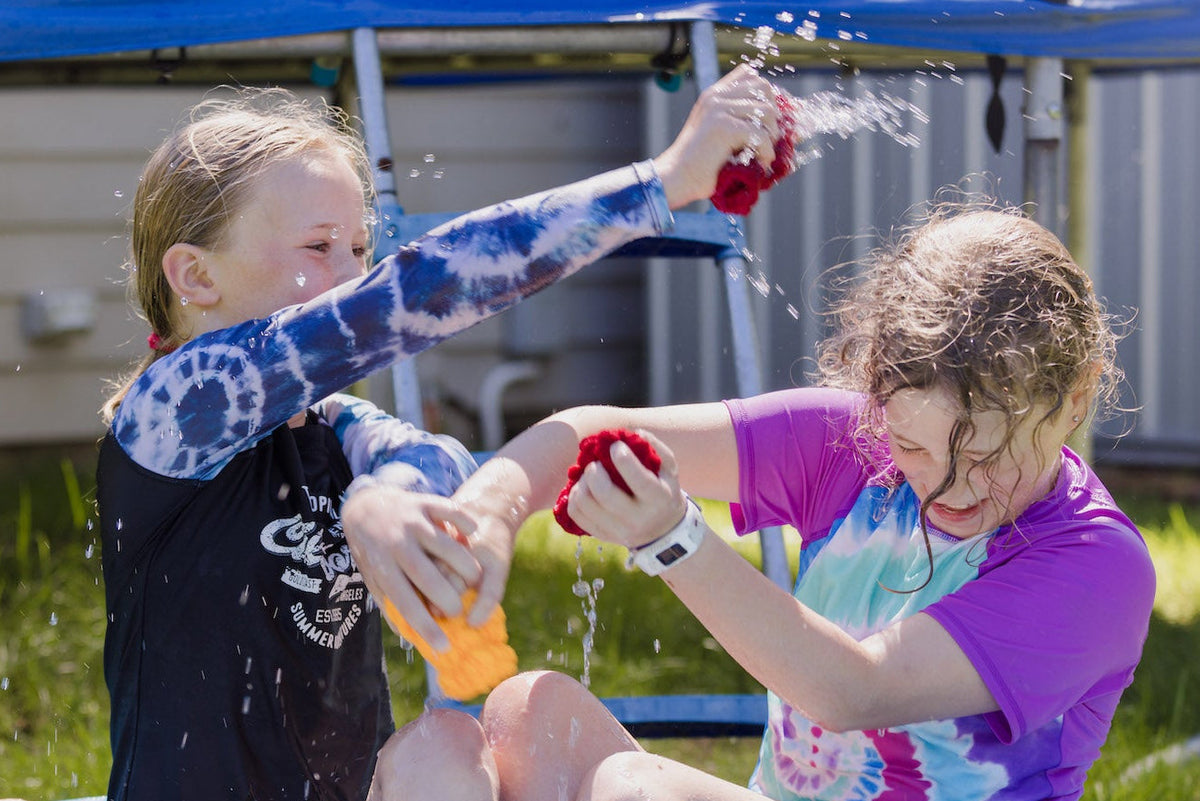 Two girls laughing and sitting in a paddling pool and playing with best eco toy for out door water fights EcoSplat Reusable Water Balloons made by New Zealand toy brand Sustained Fun