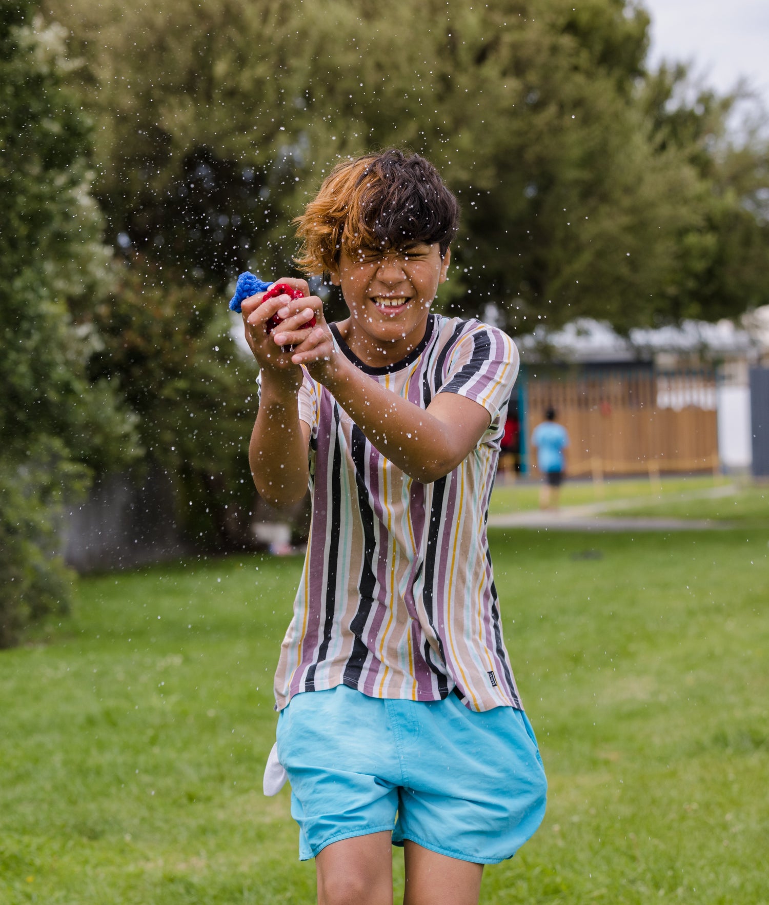 A 12 year old boy standing on grass and catching EcoSplat Reusable Water Balloon and being splashed with water. His eyes are shut and he is smiling.