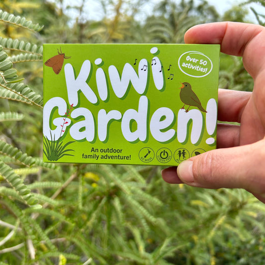 A hand holding the green eco card game box for Kiwi garden in front of Kōwhai leaves. Text on the box reads: Kiwi Garden, over 50 activities, An outdoor family adventure. There are icons saying: NZ Made, Screen free, Active, Outside. 
There are pictures of a moth, a bird singing and a harakeke flax bush with a flower spike.