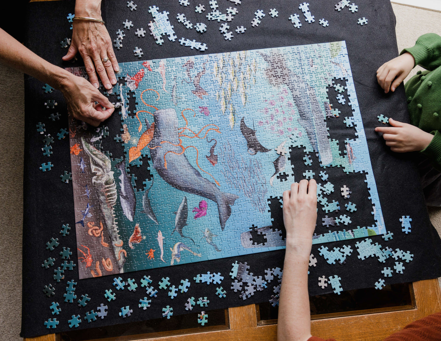A bird's-eye view of the Whale Poo and CO2 puzzle on a coffee table. The puzzle is partially completed and the hands of three people are show completing the puzzle. One is a elderly women, one is a woman and one is a young girl.