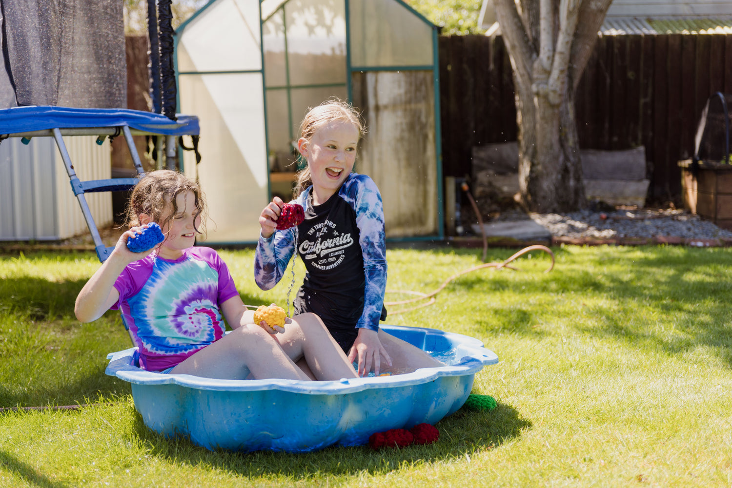 Two 11 year old girls sitting in a clam shell paddling pool in a garden, laughing and holding EcoSplat Reusable Water Balloons ready to throw them. One girl holds a red balloon and the other holds a yellow balloon and a blue balloon.