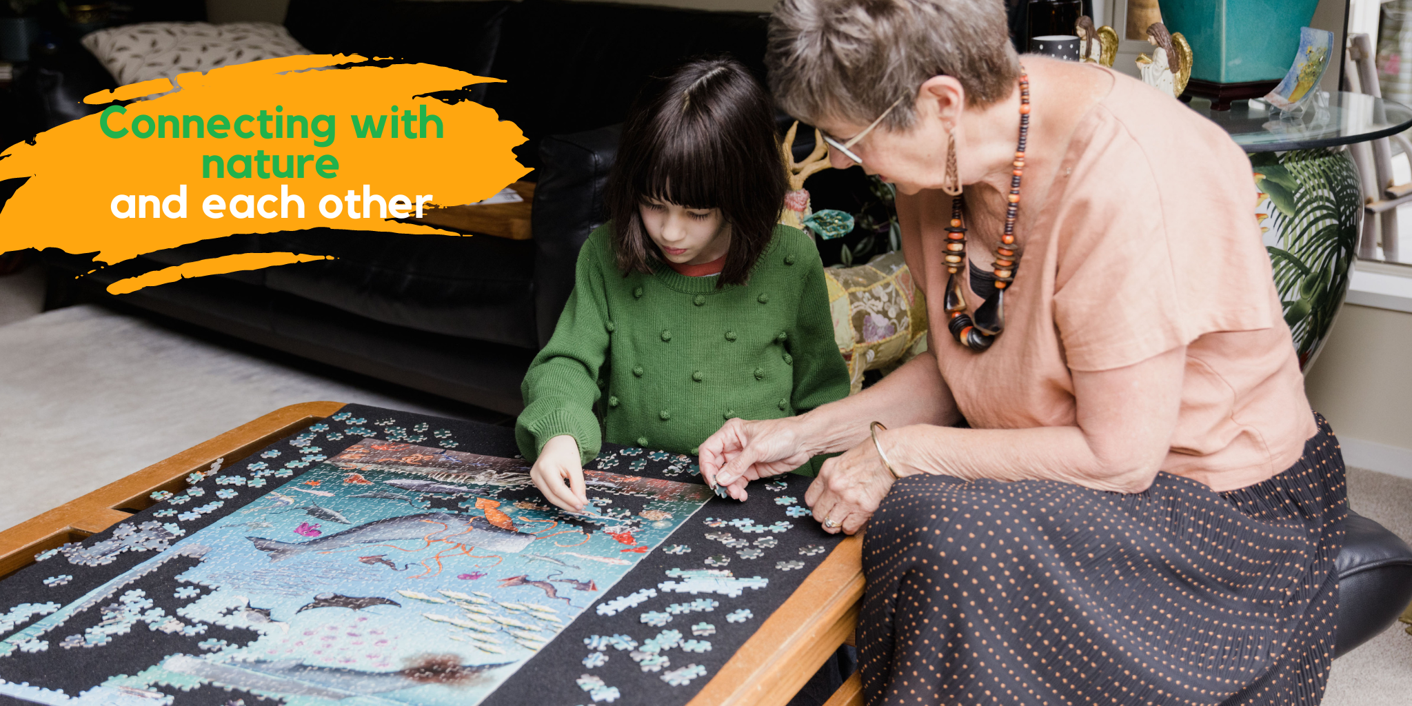A 6 year old girl and her grandmother in a living room completing a jigsaw puzzle. They are sitting next to a low coffee table. The text says "Connecting with nature and each other"